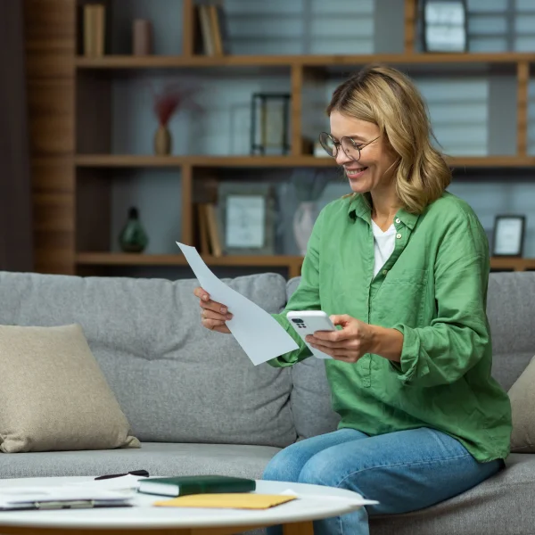 A woman sits on a couch, holding a piece of paper, appearing focused and engaged in her reading or writing.