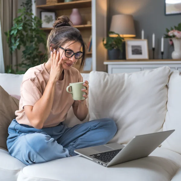 Une femme assise sur un canapé, utilisant un ordinateur portable, avec une tasse de café à la main, en situation de prêt sans emploi.