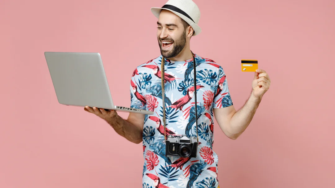 Un homme en chemise tropicale tenant une carte de crédit et un ordinateur portable, prêt pour ses voyages.